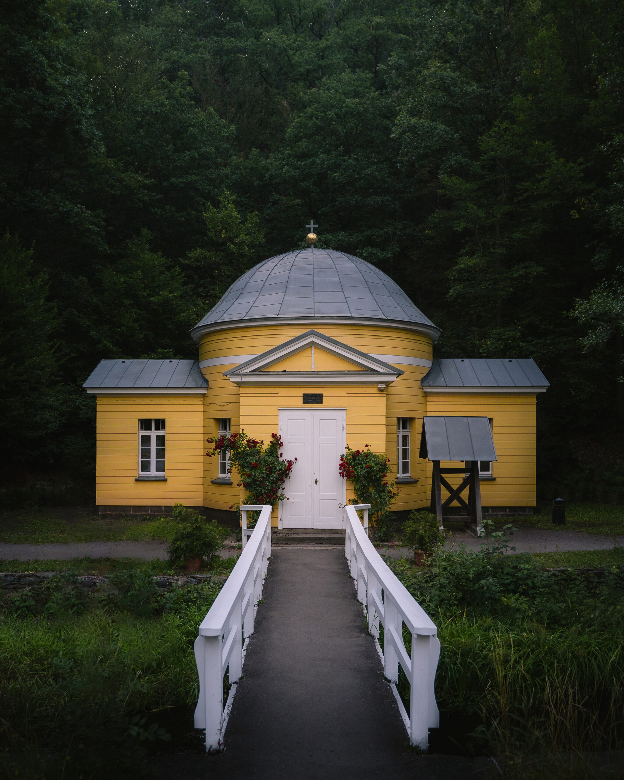 vertical front shot of a yellow christian facility with thin road and a garden in front of a forest